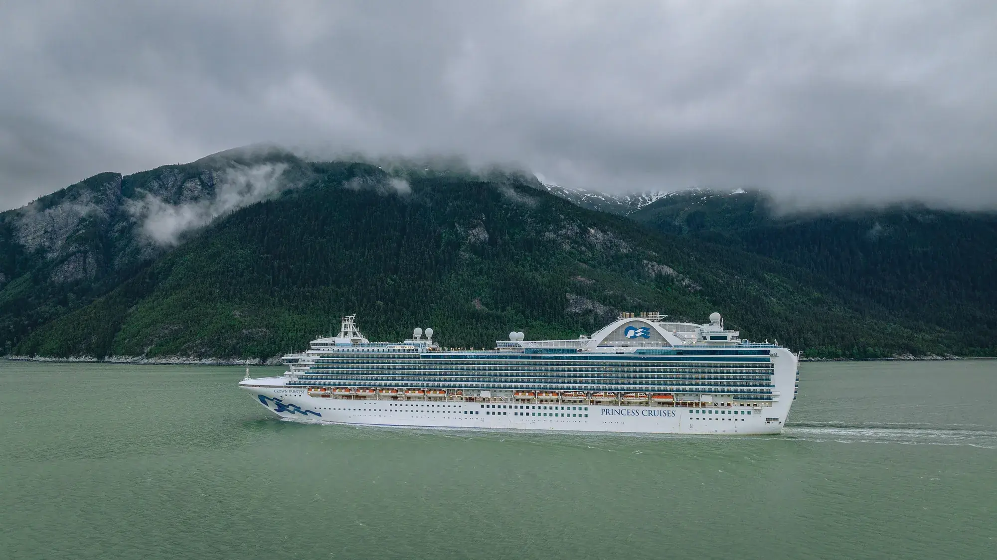 Crown Princess'in dağlık bir fiyortta yandan görünümü; pruvada Princess deniz kızı logosu